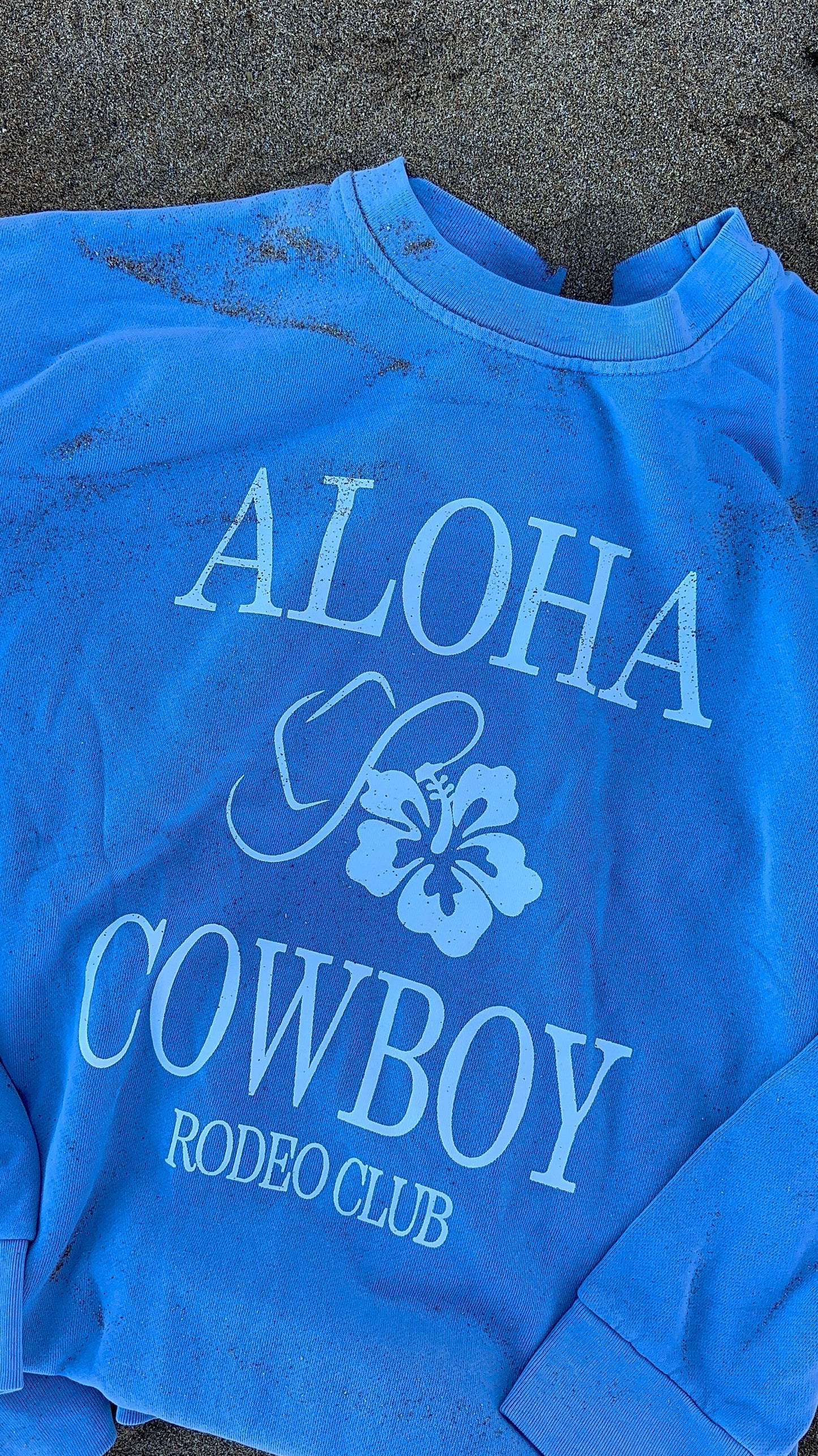 Blue crewneck with 'Aloha Cowboy Rodeo Club' text and floral design on a sandy background