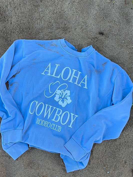 Blue sweatshirt with 'Aloha Cowboy Rodeo Club' text on a sandy surface